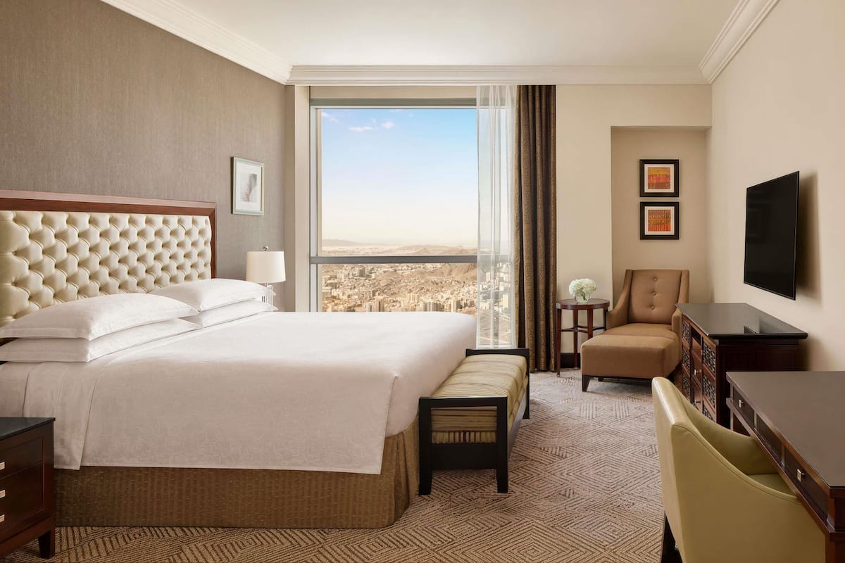 A hotel room with a large bed, a desk, a chair, a television, and a view of a cityscape.