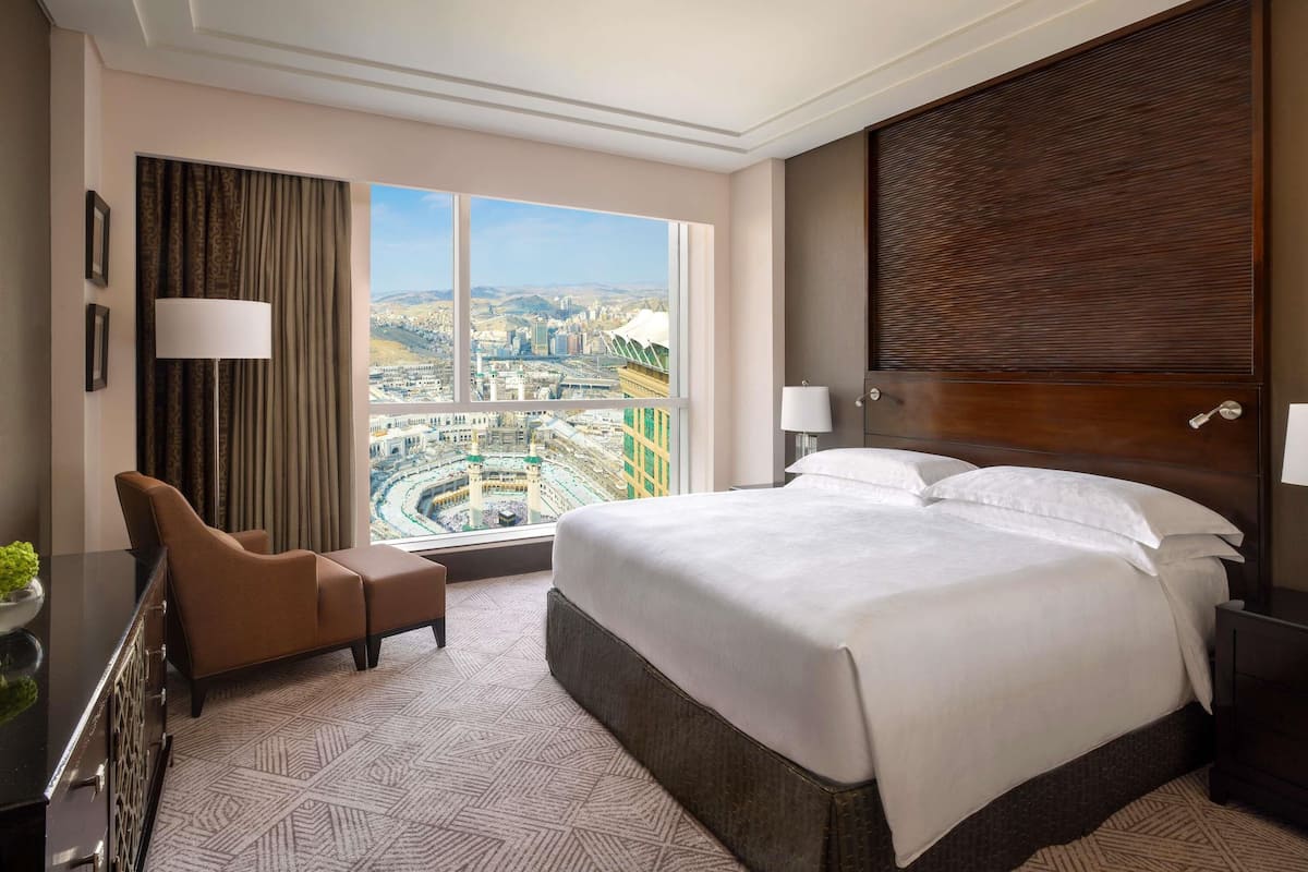 A hotel room with a large bed, a desk, a chair, a lamp, and a view of a cityscape through a large window.
