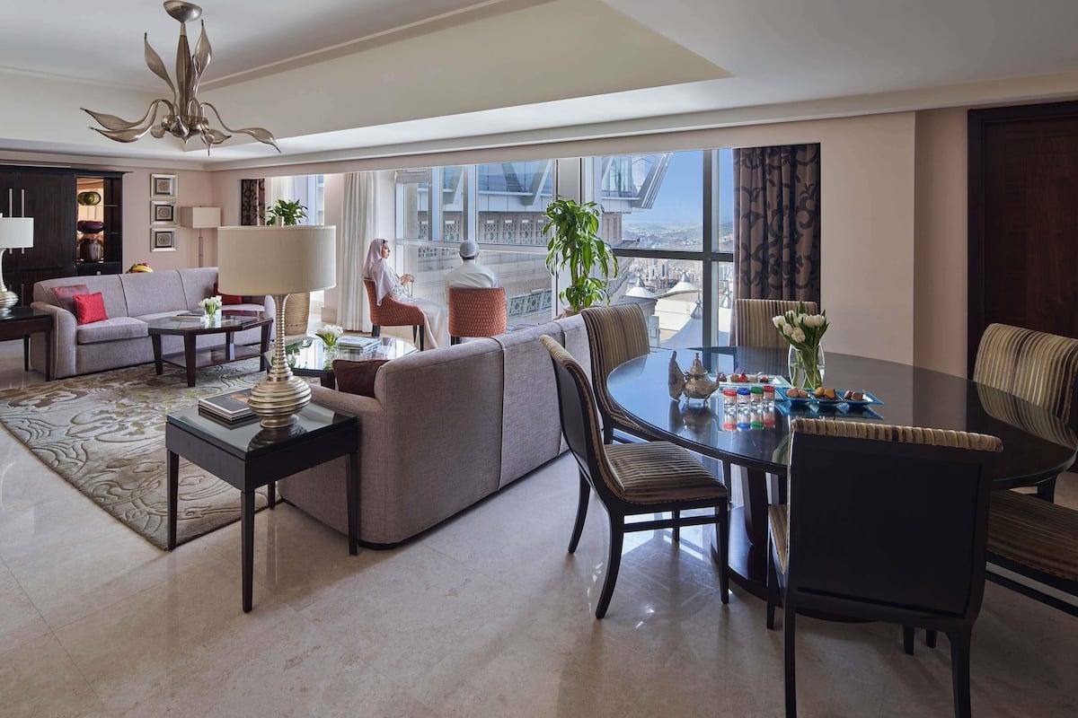A spacious living room with a sofa, coffee table, and dining table, large windows offering a city view, and a chandelier.