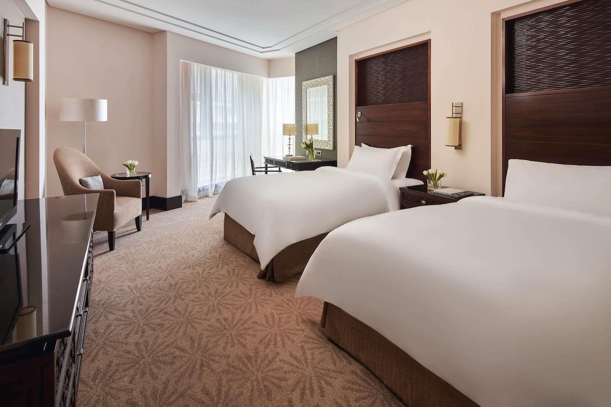 A hotel room with two beds, a desk, a chair, a TV, and a large window with curtains.