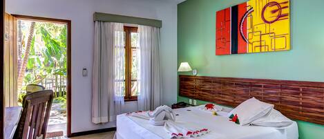 Camera d'albergo con un letto, una testiera in legno, una finestra con tende, un tavolo con una sedia e un quadro astratto dai colori vivaci appeso al muro.