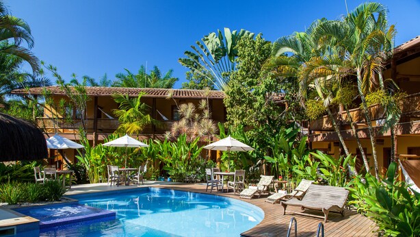 A pool area with lounge chairs and umbrellas, surrounded by tropical plants and a building with a tiled roof. - Pousada Pedra Torta (Itacaré)