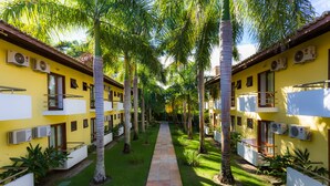 Un patio con un sendero central bordeado por palmeras y un edificio amarillo con unidades de aire acondicionado.