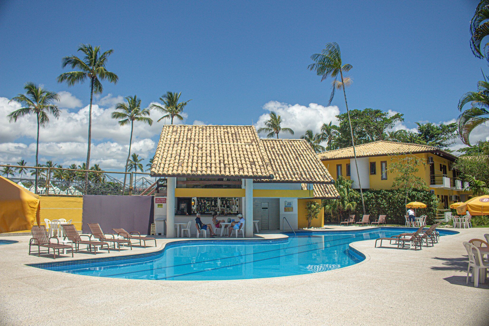 A swimming pool with lounge chairs and a bar area, surrounded by palm trees and a yellow building.