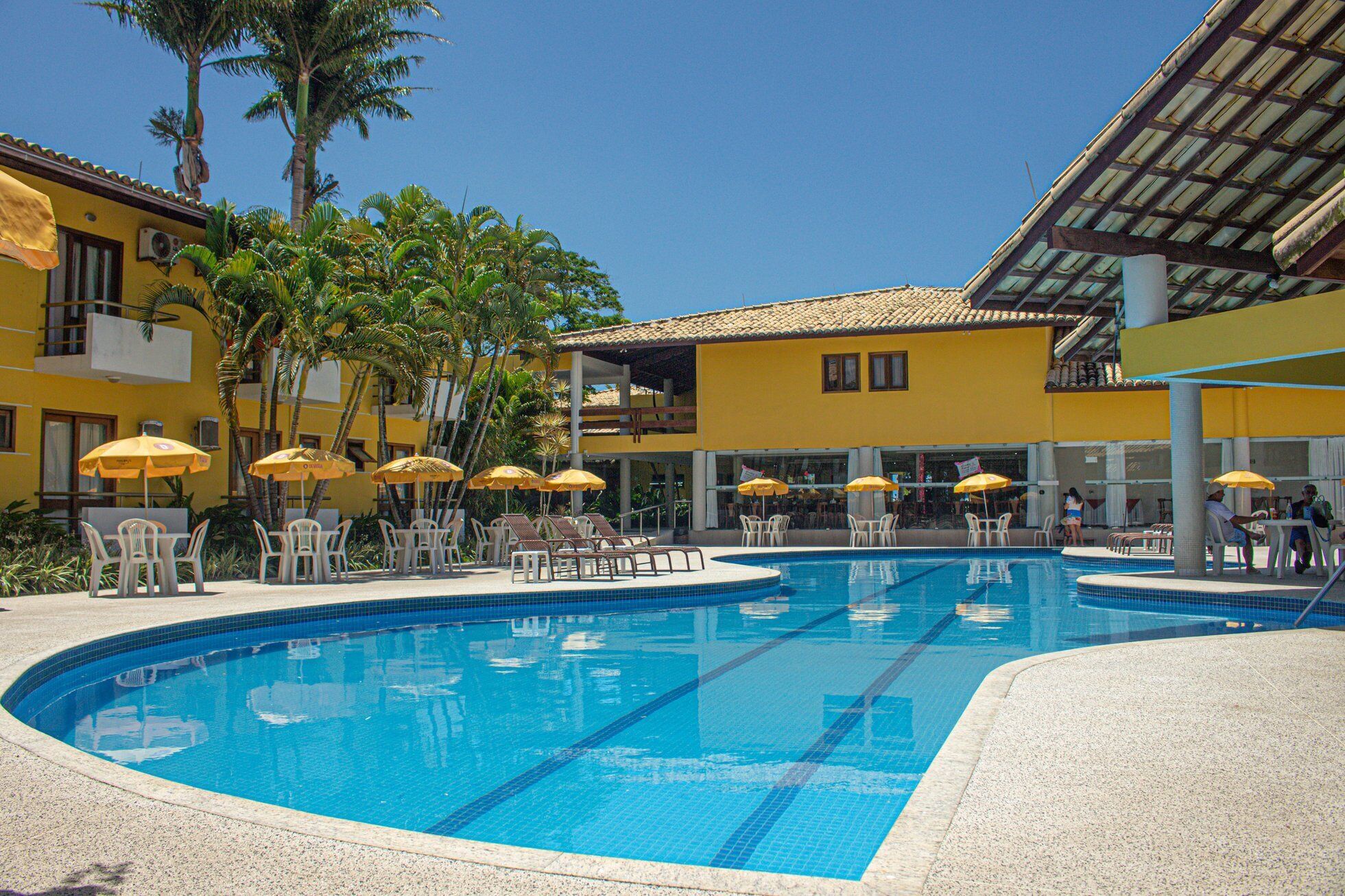 A swimming pool with lounge chairs and umbrellas, surrounded by palm trees and yellow buildings.