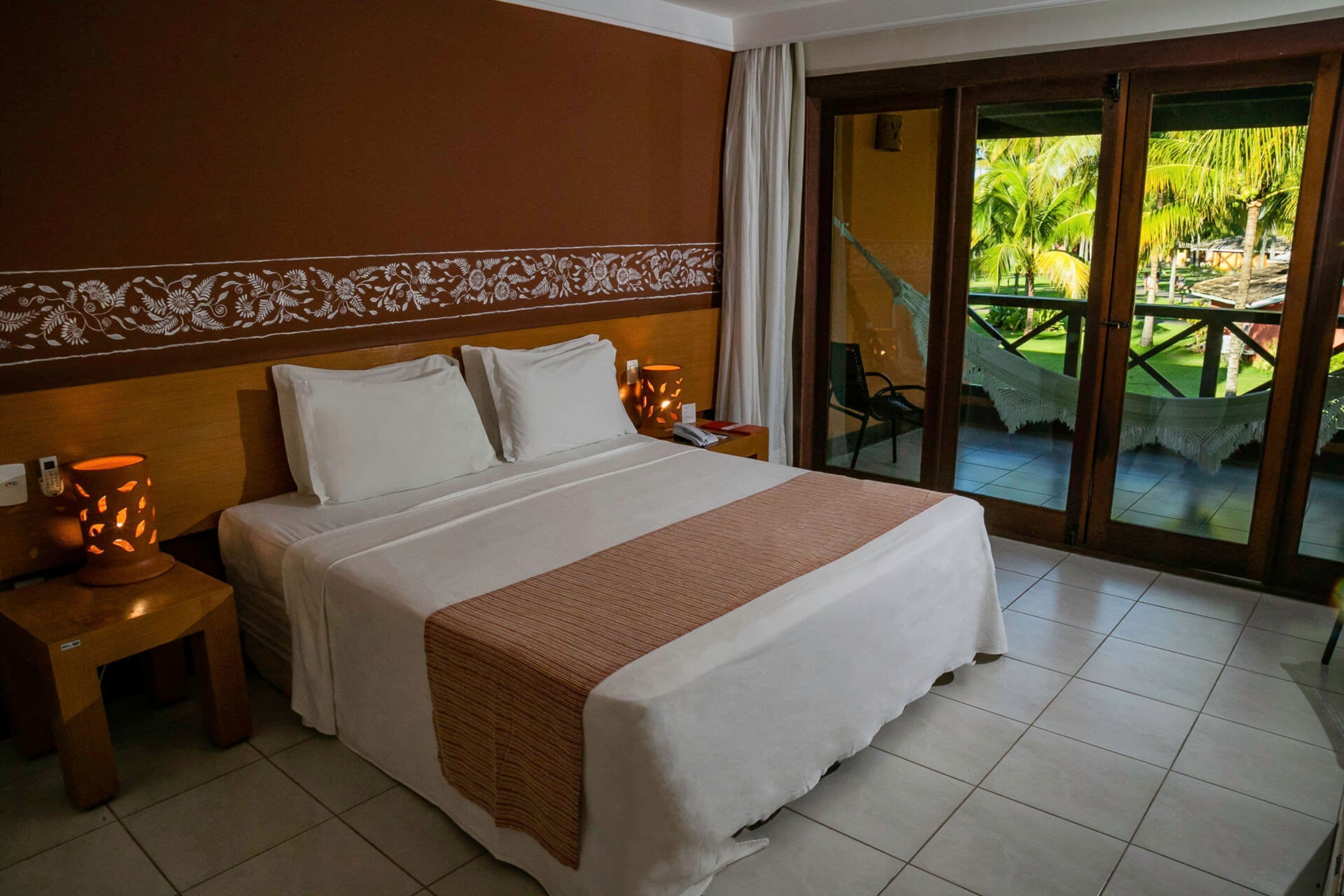 A hotel room with a large bed, a nightstand with a lamp, and a view of a palm tree and outdoor seating area through a glass door.