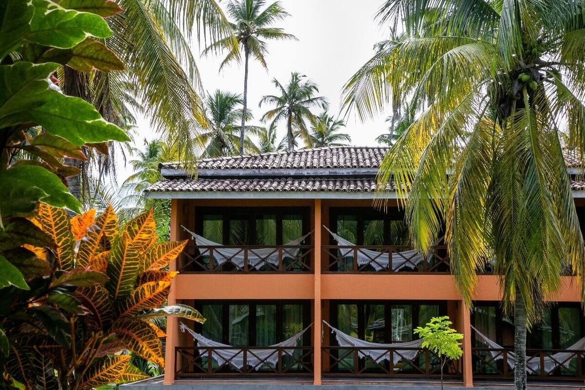 Un bâtiment à deux étages, doté d’un toit en tuiles, entouré de palmiers et de plantes tropicales.