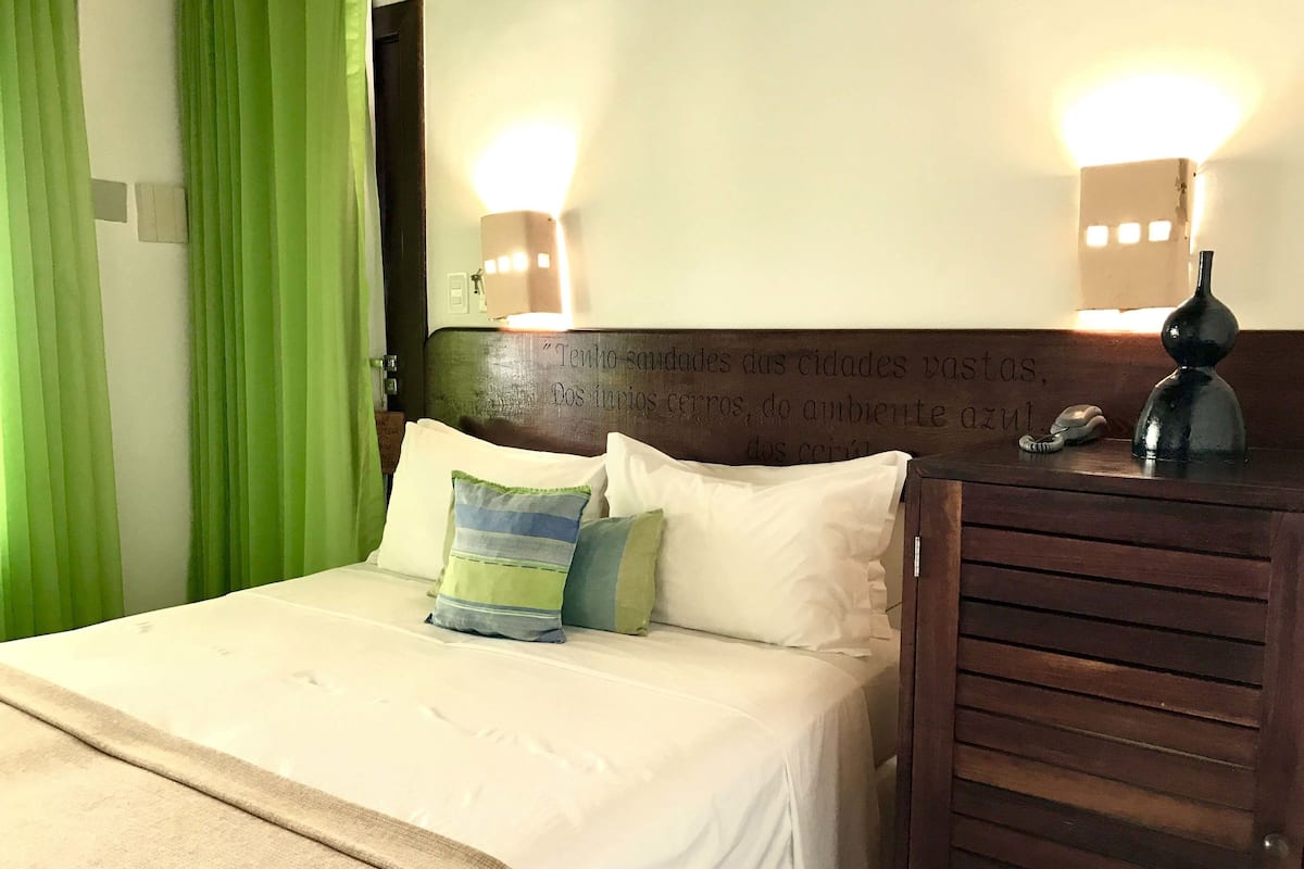 A hotel room with a bed, green curtains, a wooden headboard with a quote, and a bedside table with a lamp and a phone.