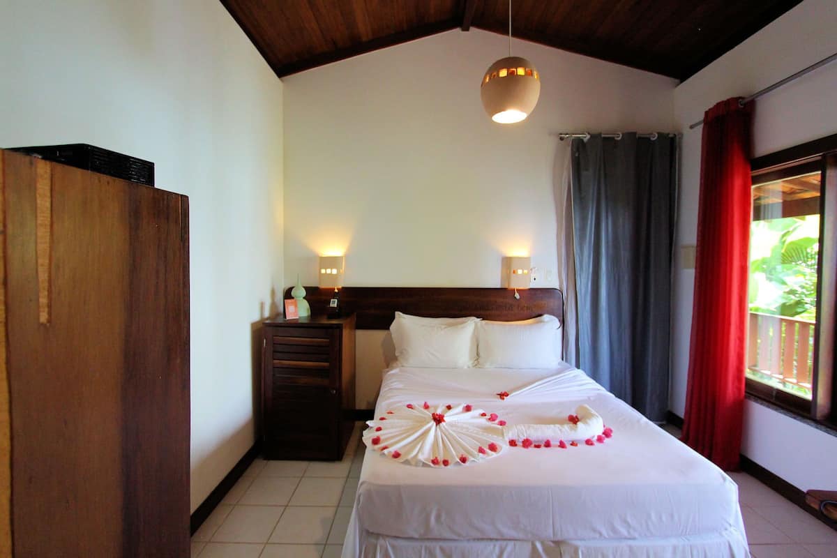 A hotel room with a bed, a wooden wardrobe, a nightstand, a wall-mounted lamp, and a window with curtains.