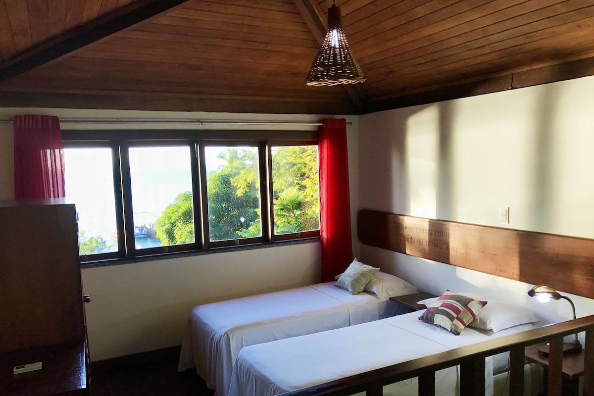 A room with two beds, a wooden headboard, a window with a view of greenery, and a hanging light fixture.