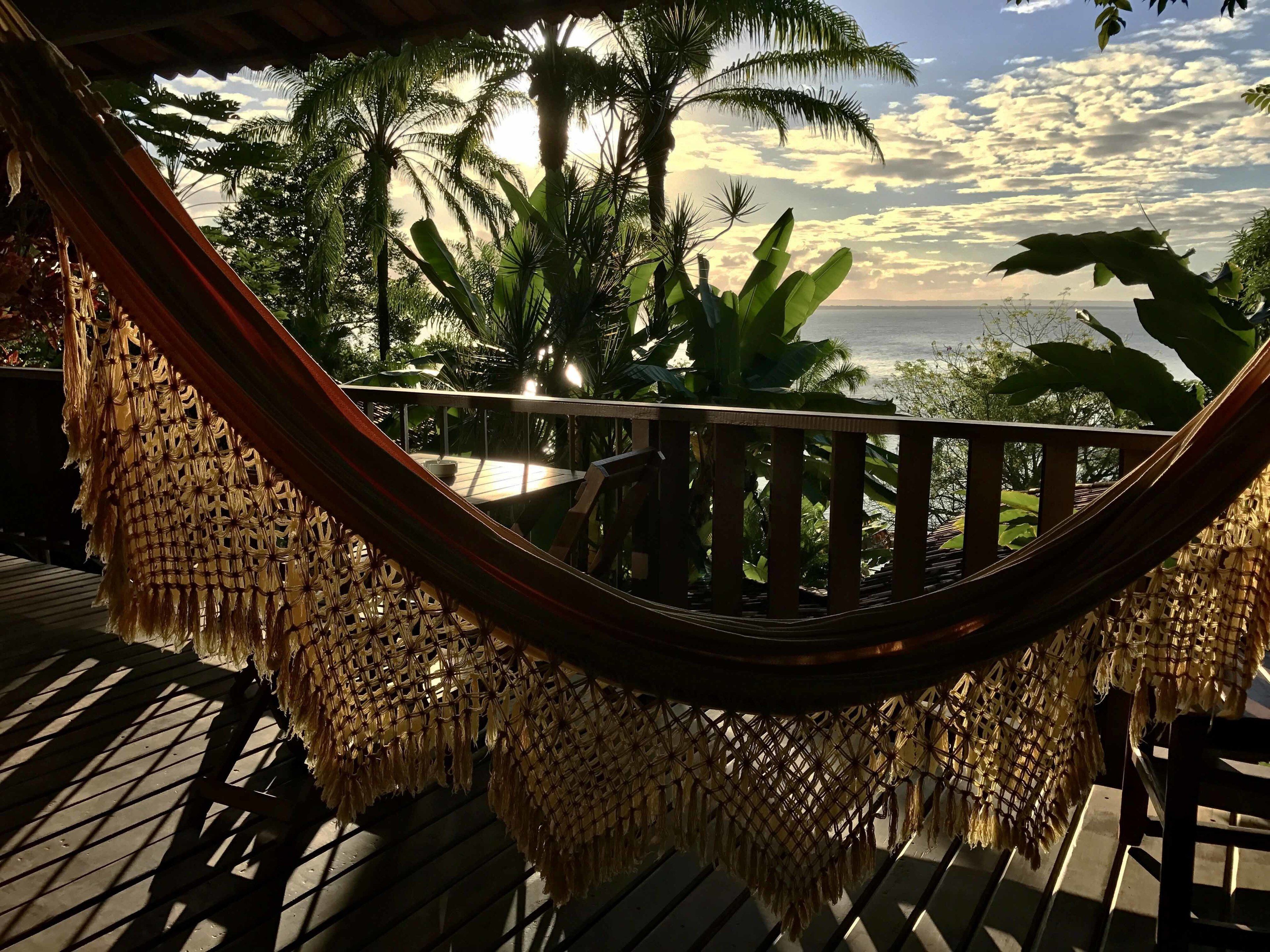 Een hangmat op een houten terras met uitzicht op palmbomen en de oceaan.