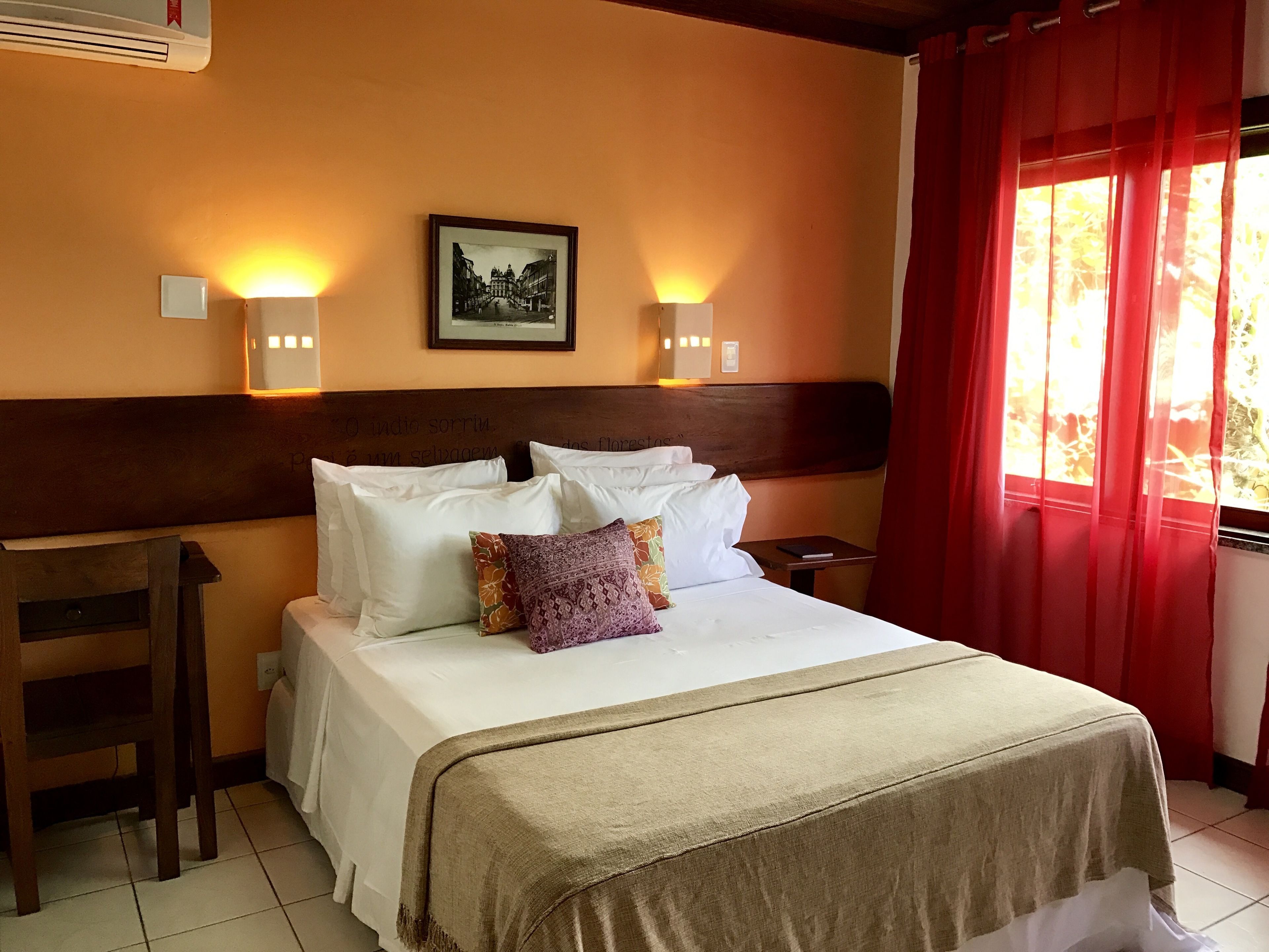 A hotel room with a bed, pillows, a headboard, a bedside table, and a window with red curtains.