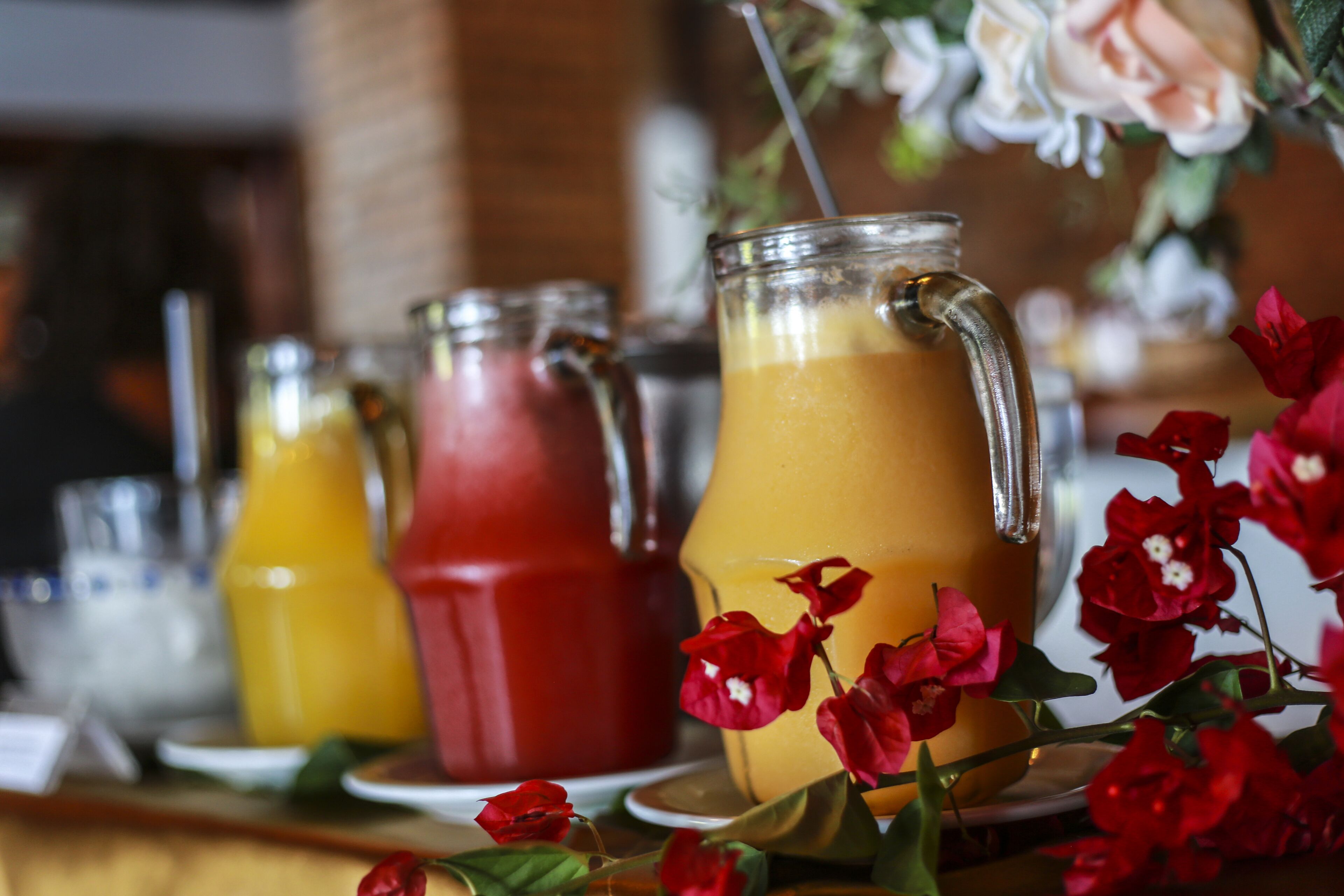 Un bar avec diverses boissons servies dans des verres et des carafes, dont une boisson jaune et une boisson rouge, et des fleurs rouges au premier plan.