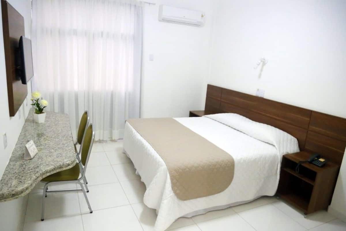 Quarto de hotel com cama, escrivaninha com cadeiras, televisão e ar condicionado.