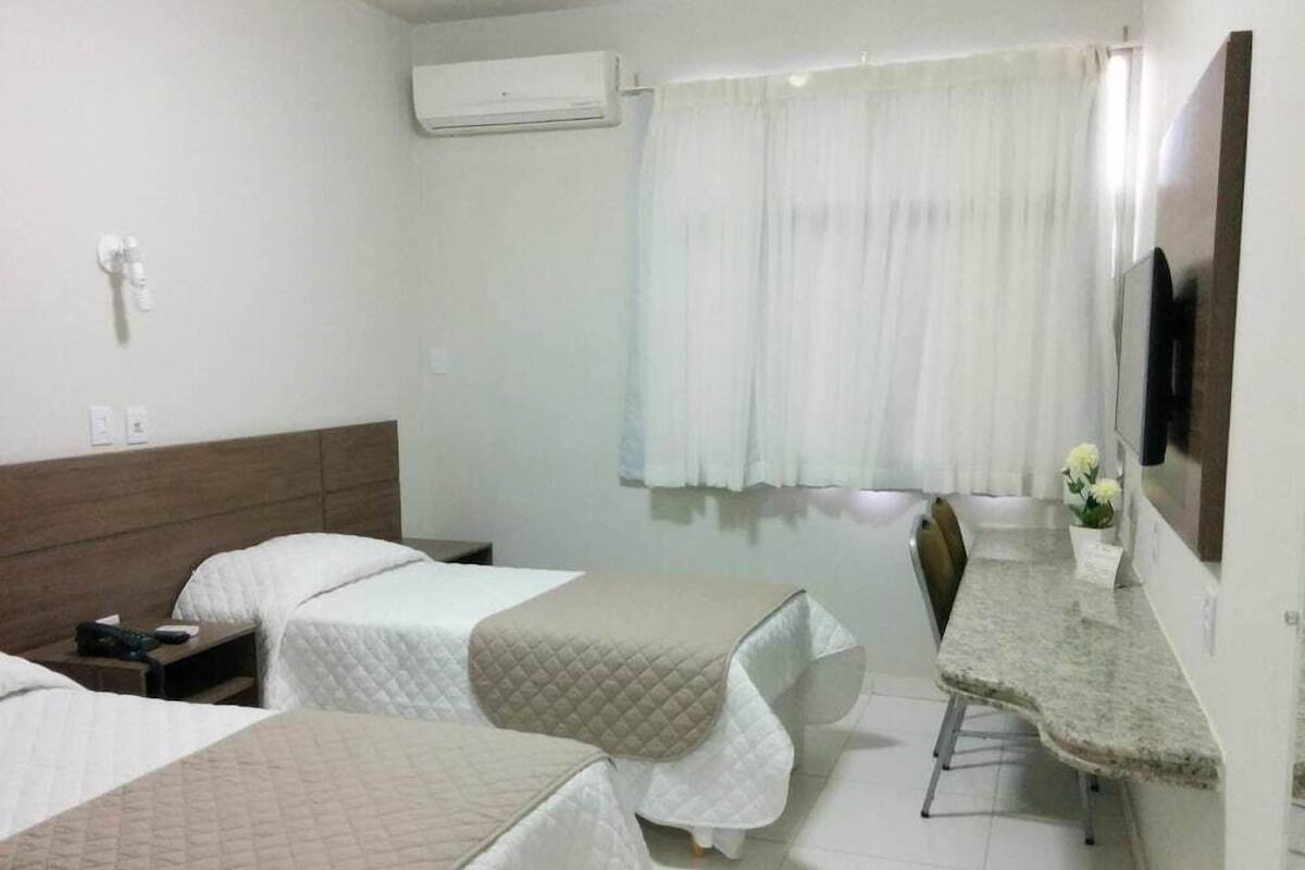 Quarto de hotel com três camas, uma escrivaninha, uma cadeira, ar condicionado e televisão presa na parede.