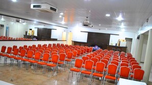 Un salón de conferencias con filas de sillas anaranjadas, una mesa blanca y una pantalla de proyección.
