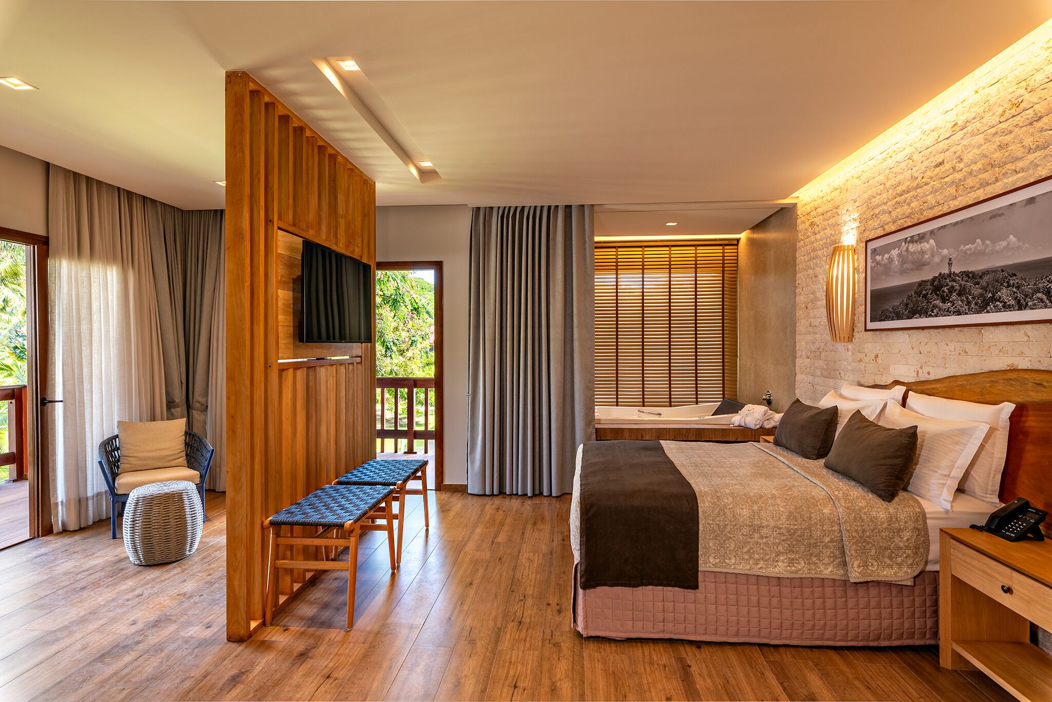 A modern bedroom with a large bed, wooden furniture, a TV, and a view of greenery outside.