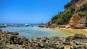 Una playa rocosa con agua cristalina, una zona arenosa y un acantilado con vegetación.