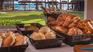 Una mesa de buffet con diferentes panes y pasteles, con vistas a una playa que tiene sombrillas y sillas de descanso.