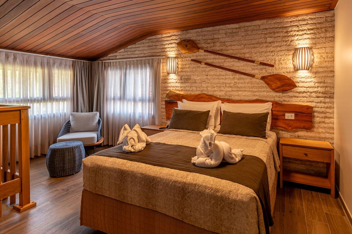 Een knus slaapkamer met een houten plafond, een bakstenen muur en een bed met houten hoofdeinde. Er staan twee nachtkastjes en een stoel.