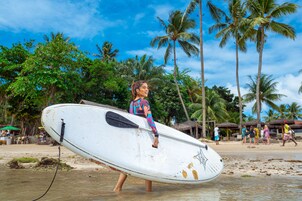 En person bærer en surfbræt på en strand med palmer og strandhytter i baggrunden.