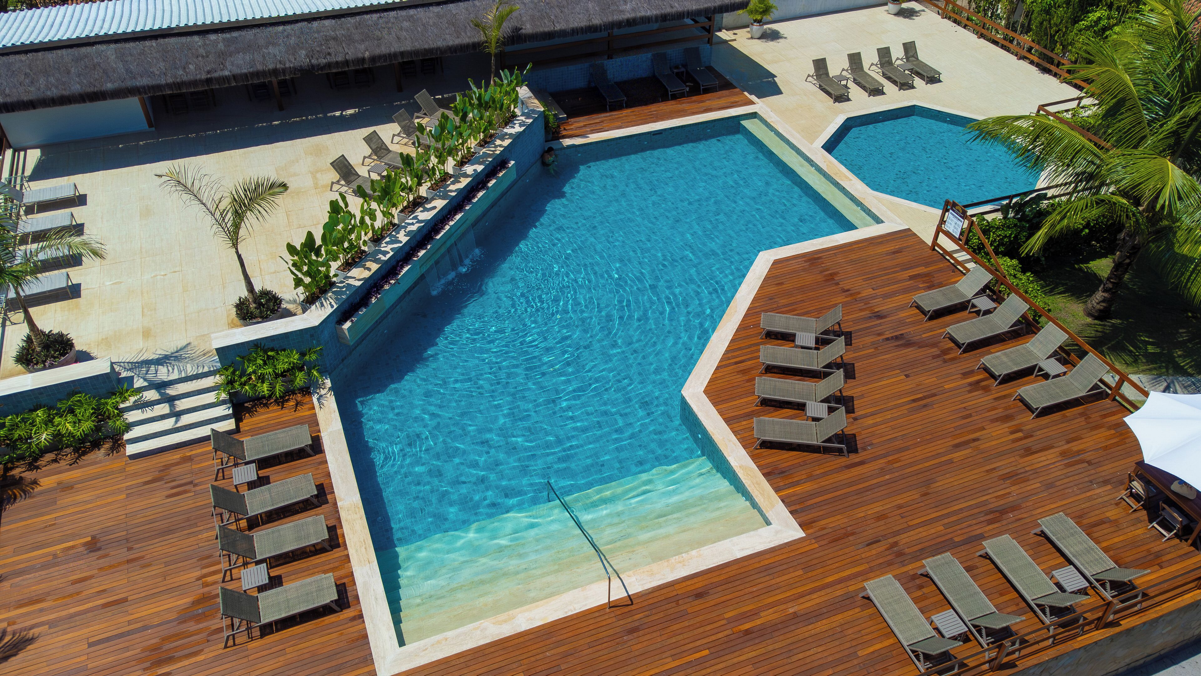 Una gran piscina rectangular con un deck de madera a su alrededor, con varias sillas de descanso y una piscina circular más pequeña.