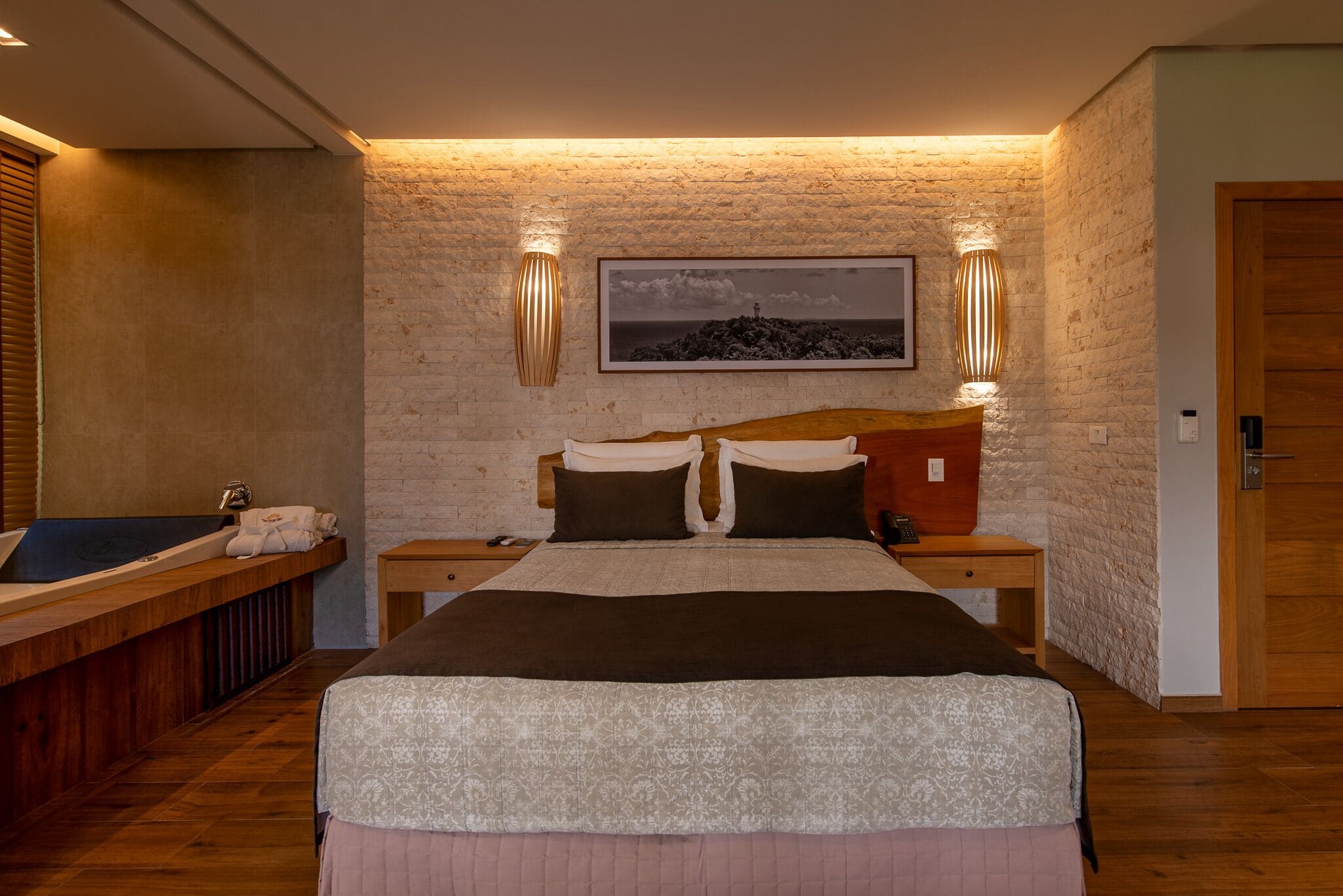 Quarto com uma cama grande, cabeceira de madeira, duas mesas de cabeceira, um quadro na parede e um banco de madeira com uma toalha.
