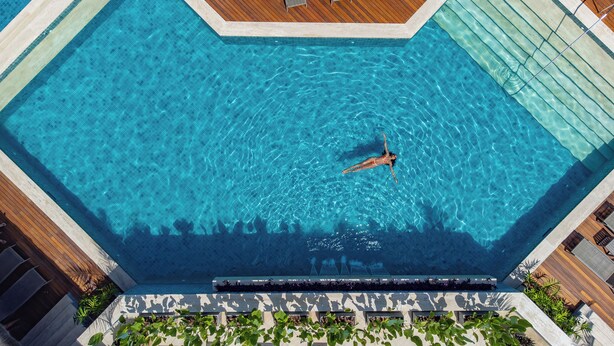 Vista aérea de una piscina con una persona nadando, rodeada de un muelle de madera y vegetación.