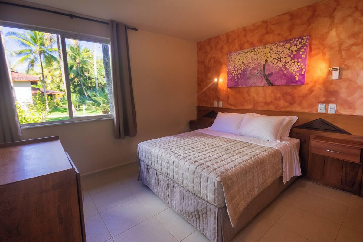 Een slaapkamer met een groot bed, een houten nachtkastje, een dressoir en een raam met uitzicht op palmbomen.