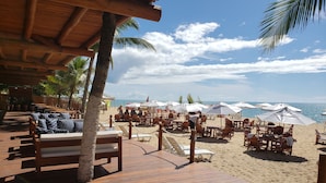 A beachside restaurant with outdoor seating, palm trees, and a view of the ocean.