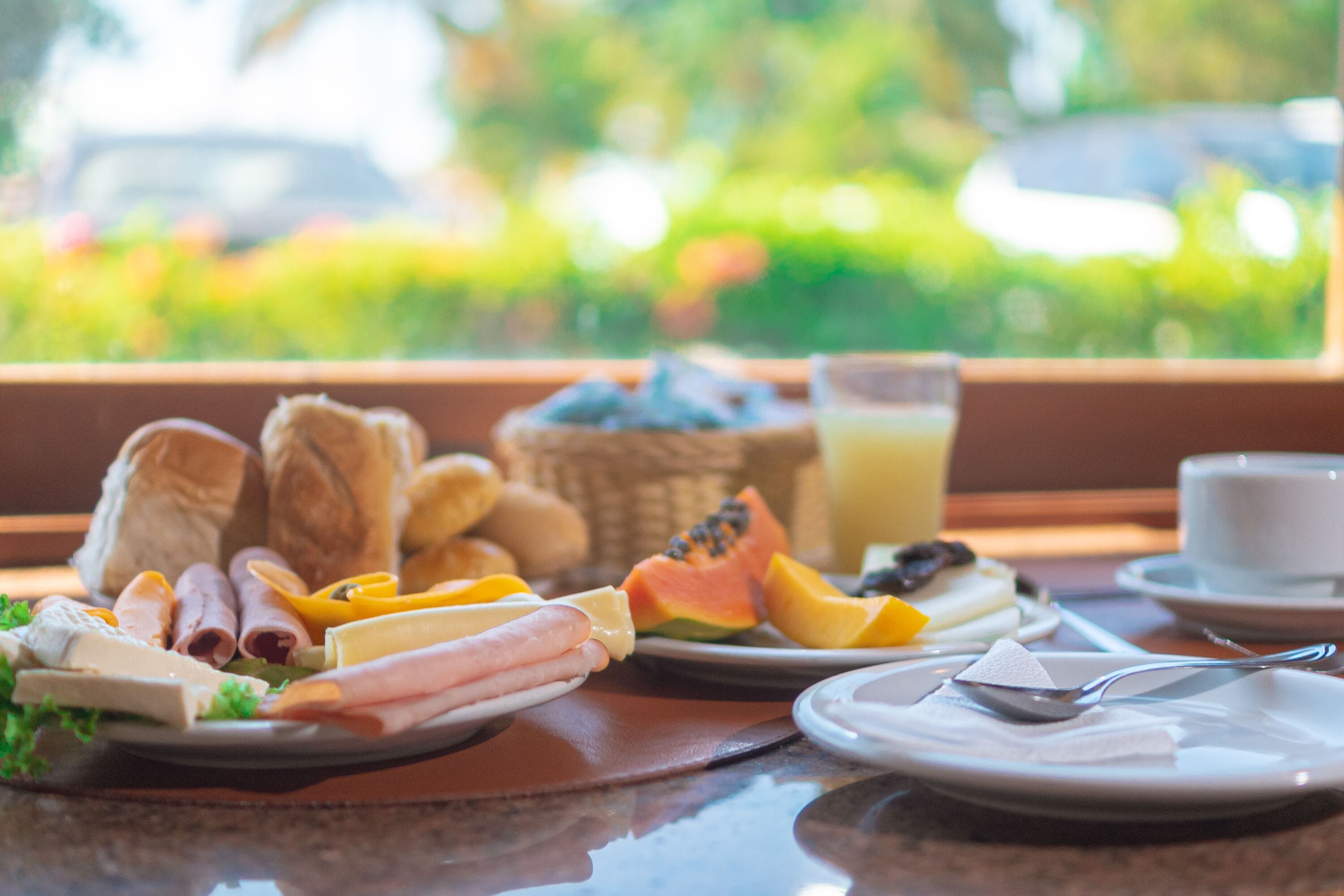 A breakfast table with various foods and a glass of juice, set against a window with a view of greenery.