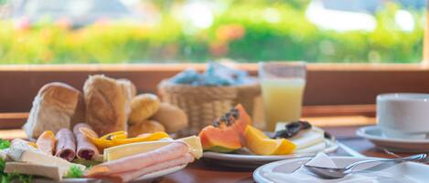 A breakfast table with various foods and a glass of juice, set against a window with a view of greenery.
