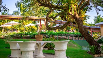 Un jardín con una mesa central rodeada de sillas, un área de estar cubierta con sombrillas y abundante vegetación.