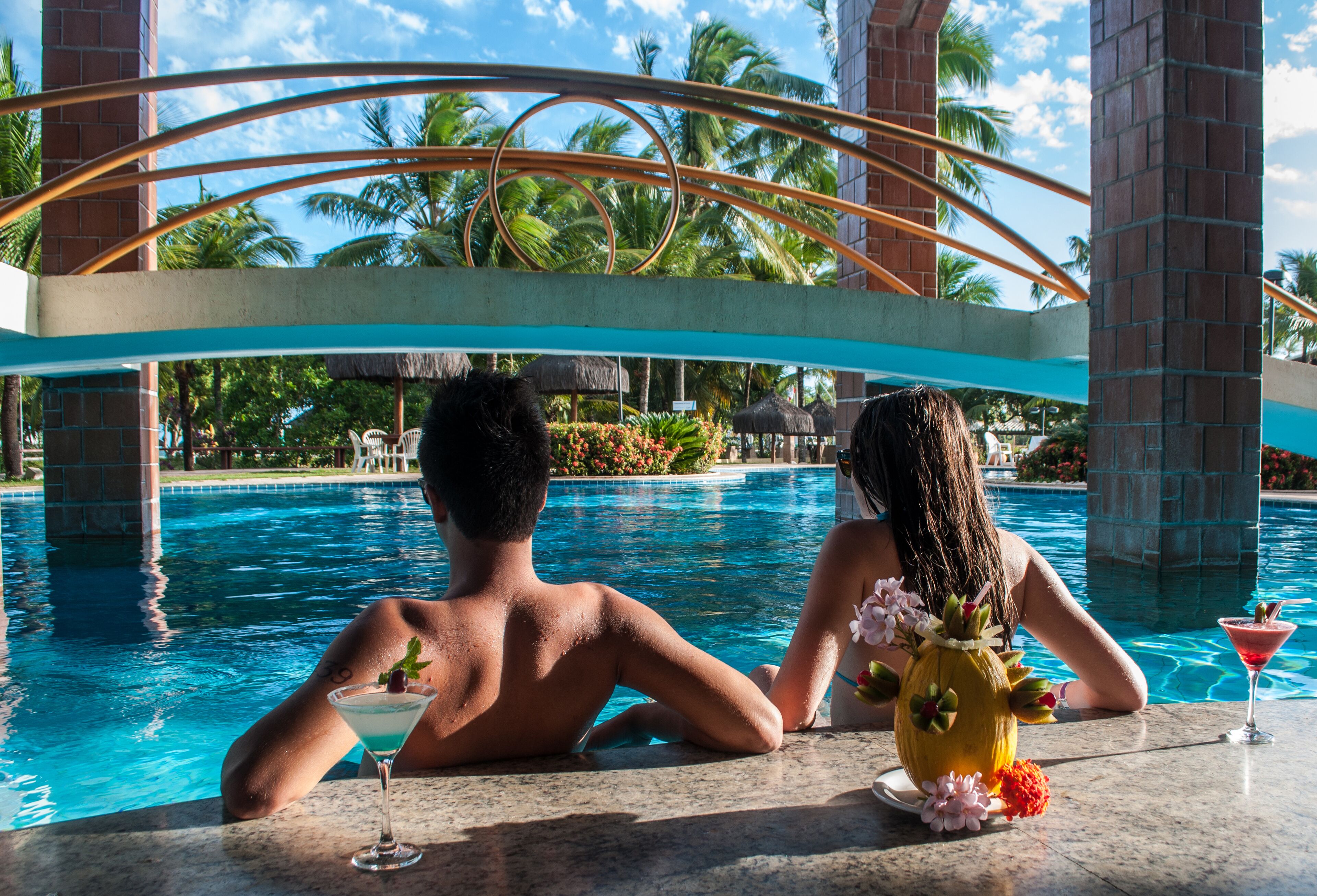 A couple relaxing by a pool with a tropical backdrop, including palm trees and a thatched-roof structure.