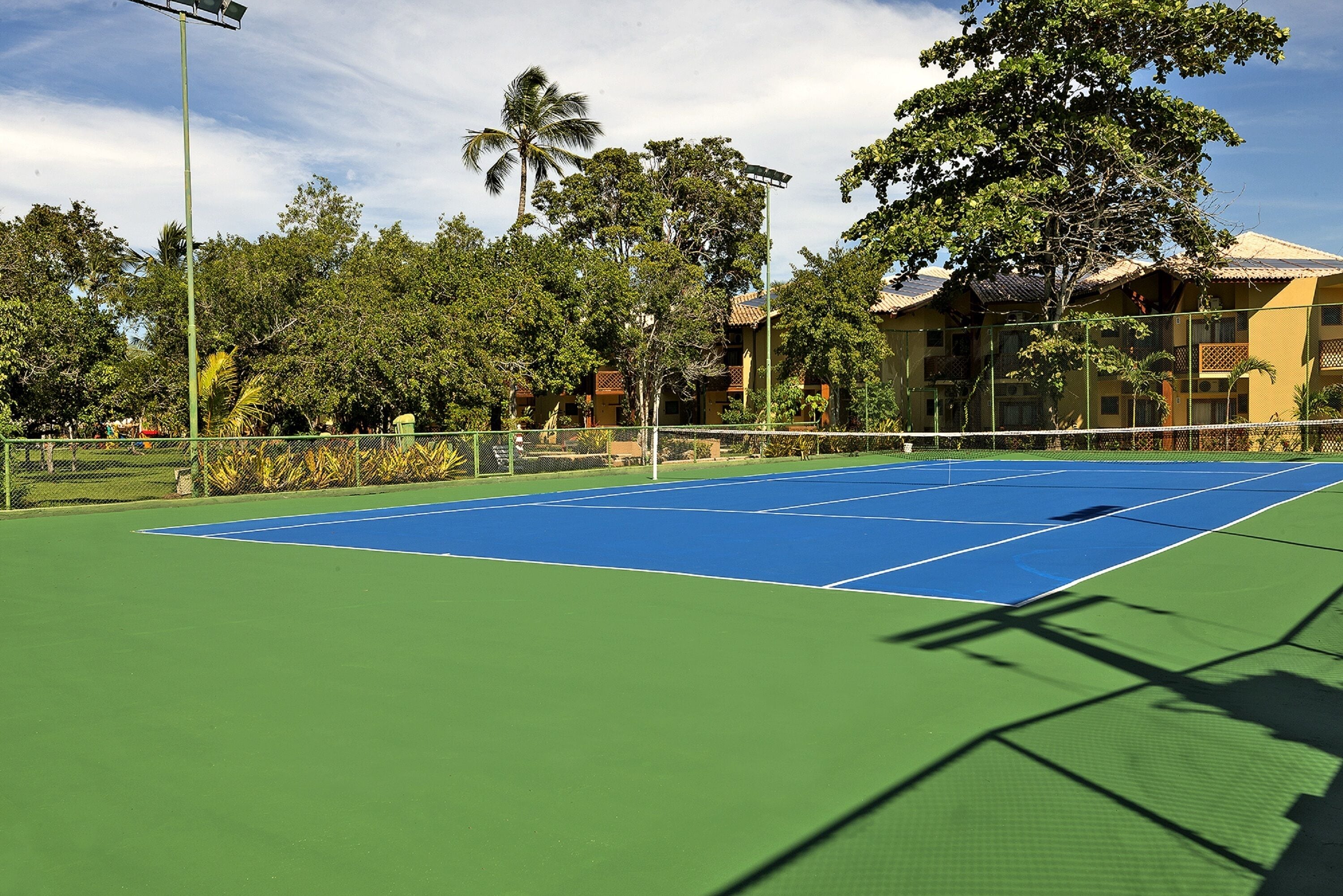 A tennis court with a green and blue surface, surrounded by trees and a building with balconies.