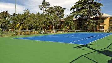 A tennis court with a green and blue surface, surrounded by trees and a building with balconies.