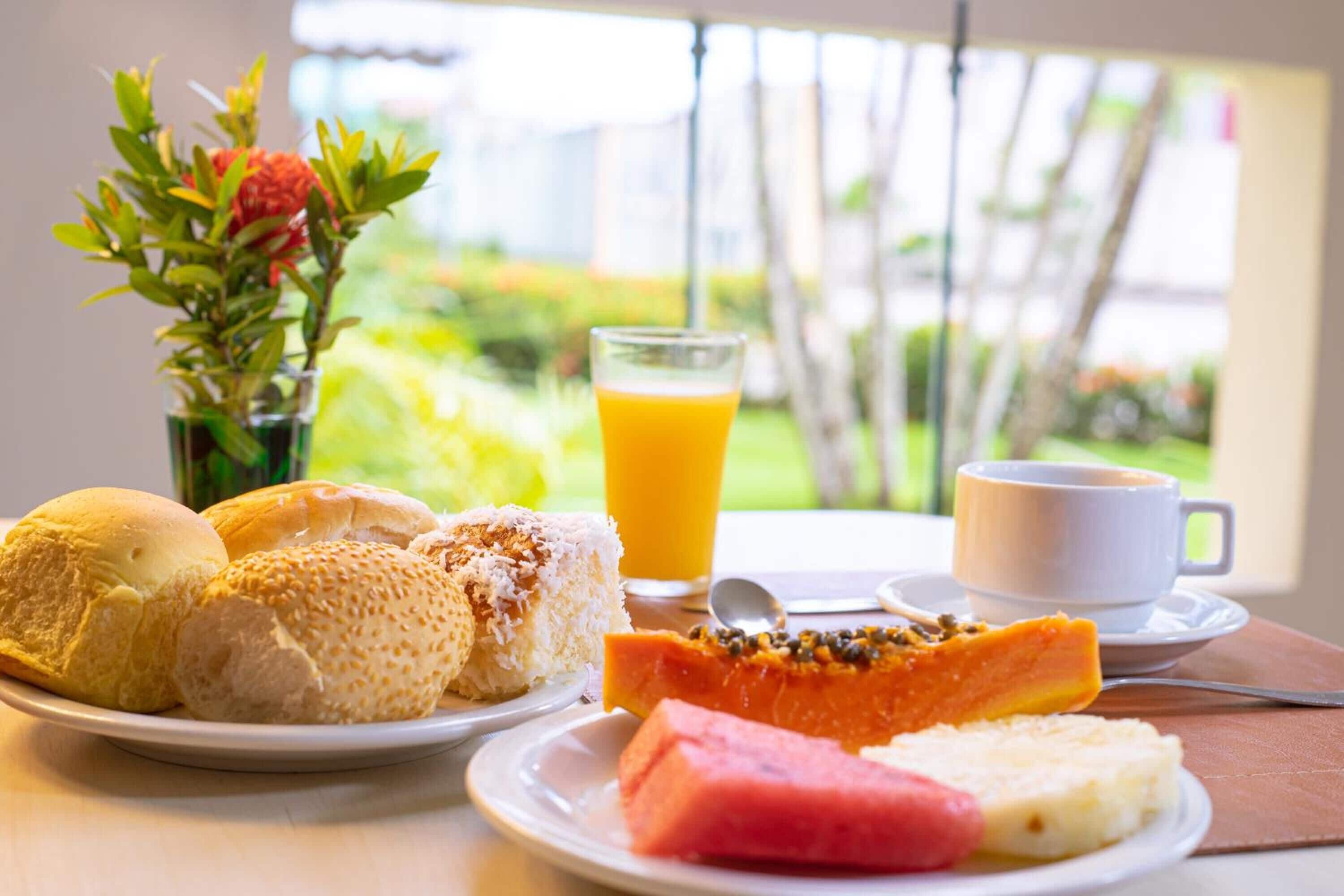A breakfast setting with bread rolls, a slice of watermelon, a piece of cake, a glass of orange juice, and a cup of coffee on a table with a view of greenery outside.