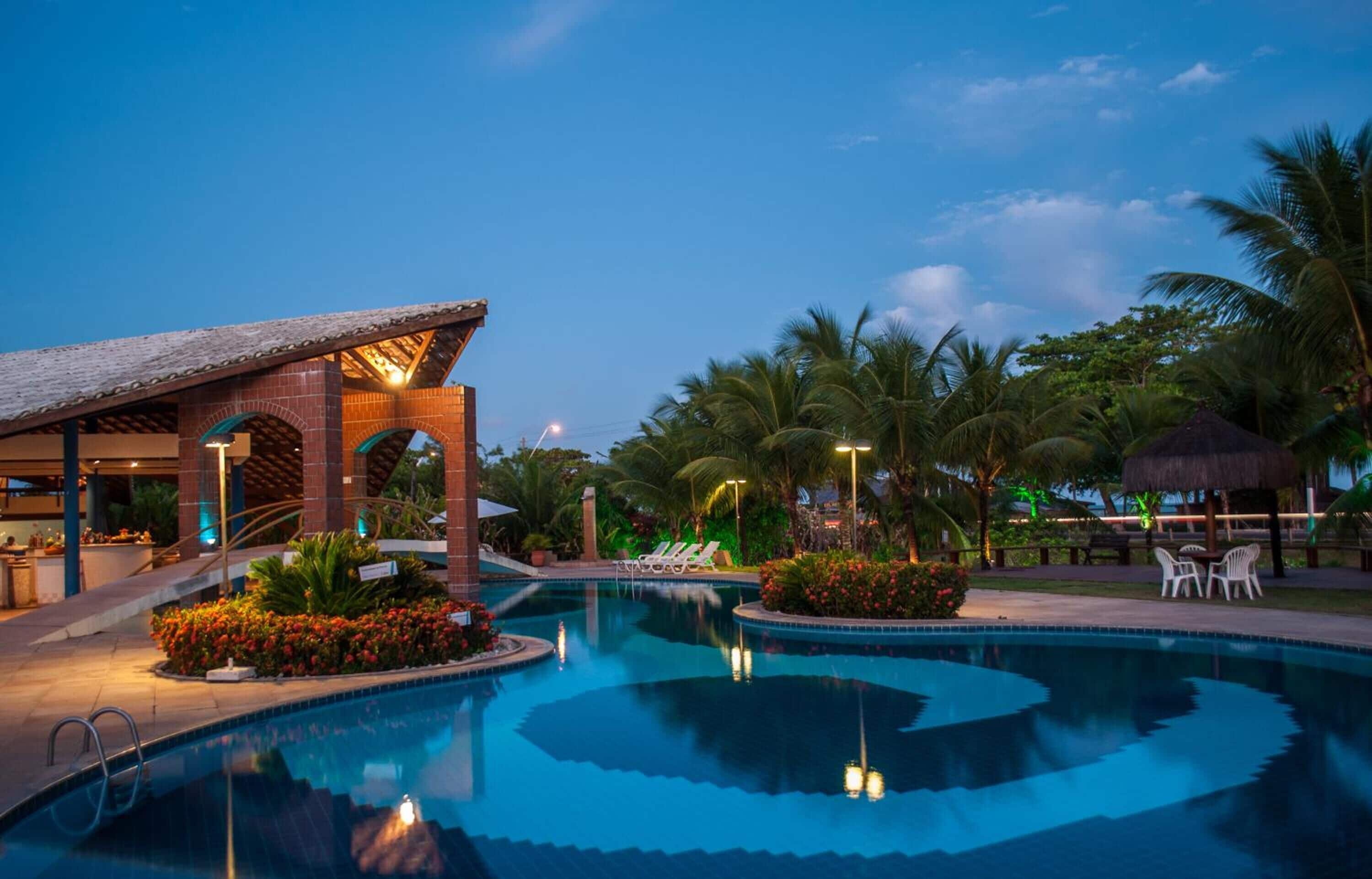 A swimming pool with a thatched-roof structure, palm trees, and lounge chairs.