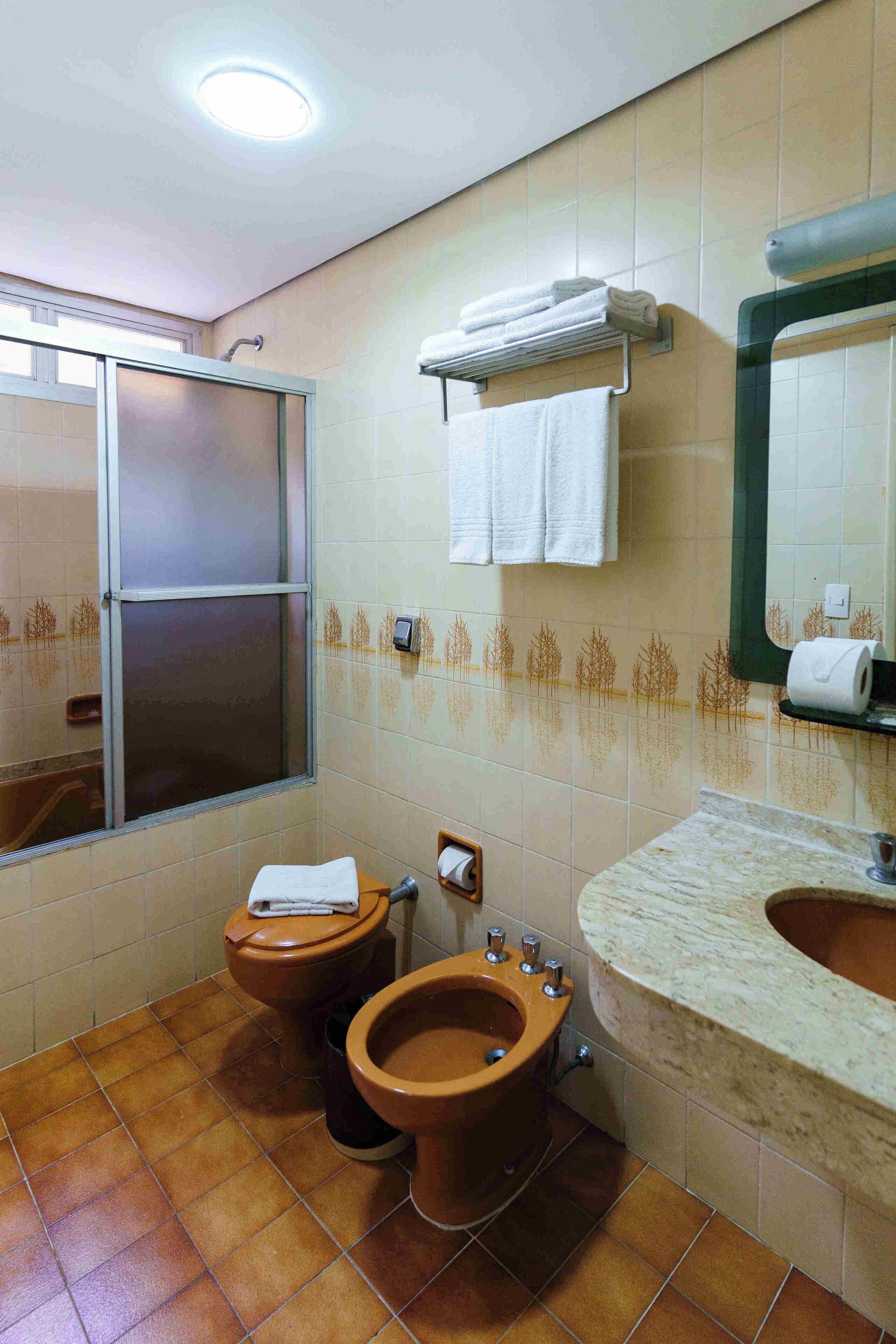 A bathroom with a toilet, bidet, and sink, tiled in brown and beige, with a window and a towel rack.