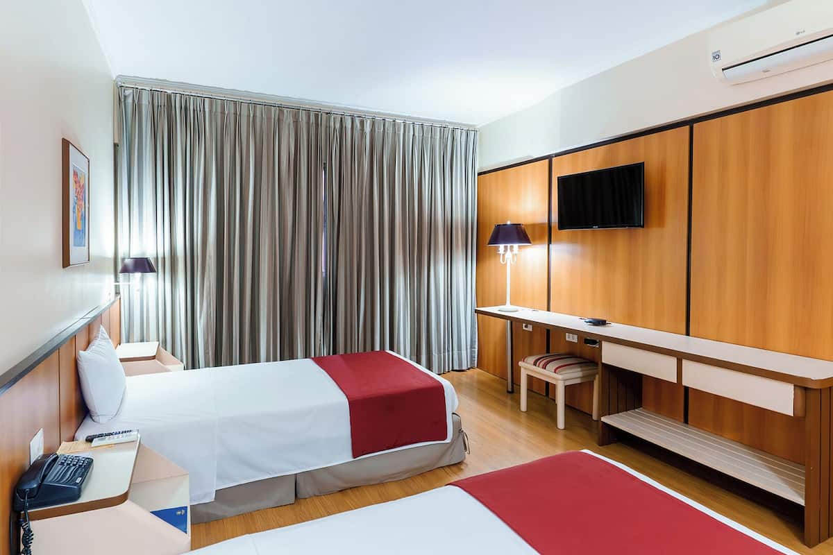 Ein Hotelzimmer mit zwei Betten, einem Schreibtisch, einem Stuhl, einem Fernseher und einem Vorhangfenster.