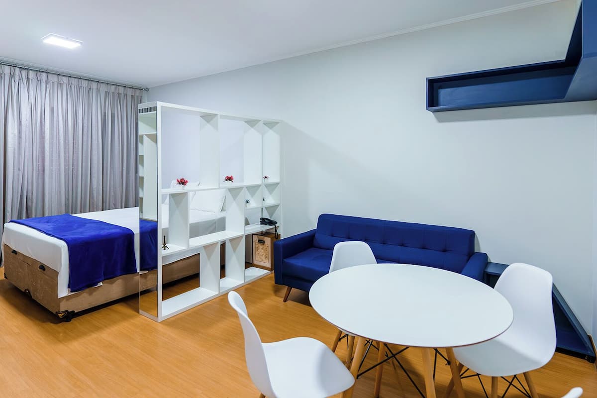 A hotel room with a bed, a sofa, a small table, and a chair. There is a white partition with shelves and a small cabinet.