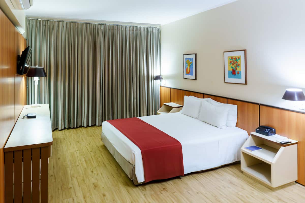 A hotel room with a large bed, wooden floor, a desk with a lamp, a nightstand, and two framed pictures on the wall.