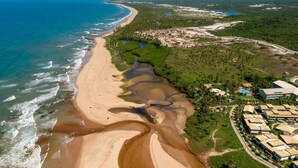An aerial view of a coastal area with a sandy beach, a river, and a residential area with buildings. - Hotel Resort Costa Dos Coqueiros (Mata de Sao Joao)