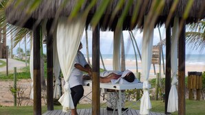 A wooden deck with a massage table under a thatched roof, a person receiving a massage, and a beach view in the background. - Hotel Resort Costa Dos Coqueiros (Mata de Sao Joao)