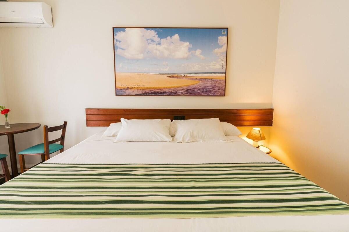 Une chambre d’hôtel avec un lit, une tête de lit en bois, une table de chevet avec une lampe, un tableau représentant une scène de plage, et une petite table avec une chaise.
