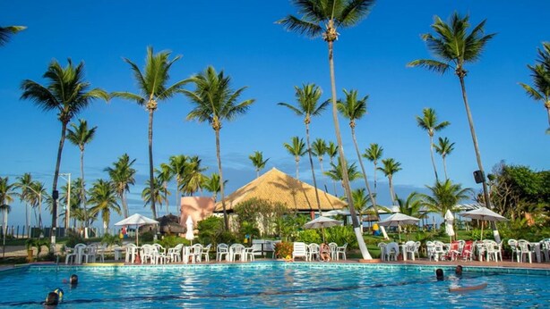 A pool area with palm trees, lounge chairs, and a thatched-roof building. - Hotel Resort Costa Dos Coqueiros (Mata de Sao Joao)