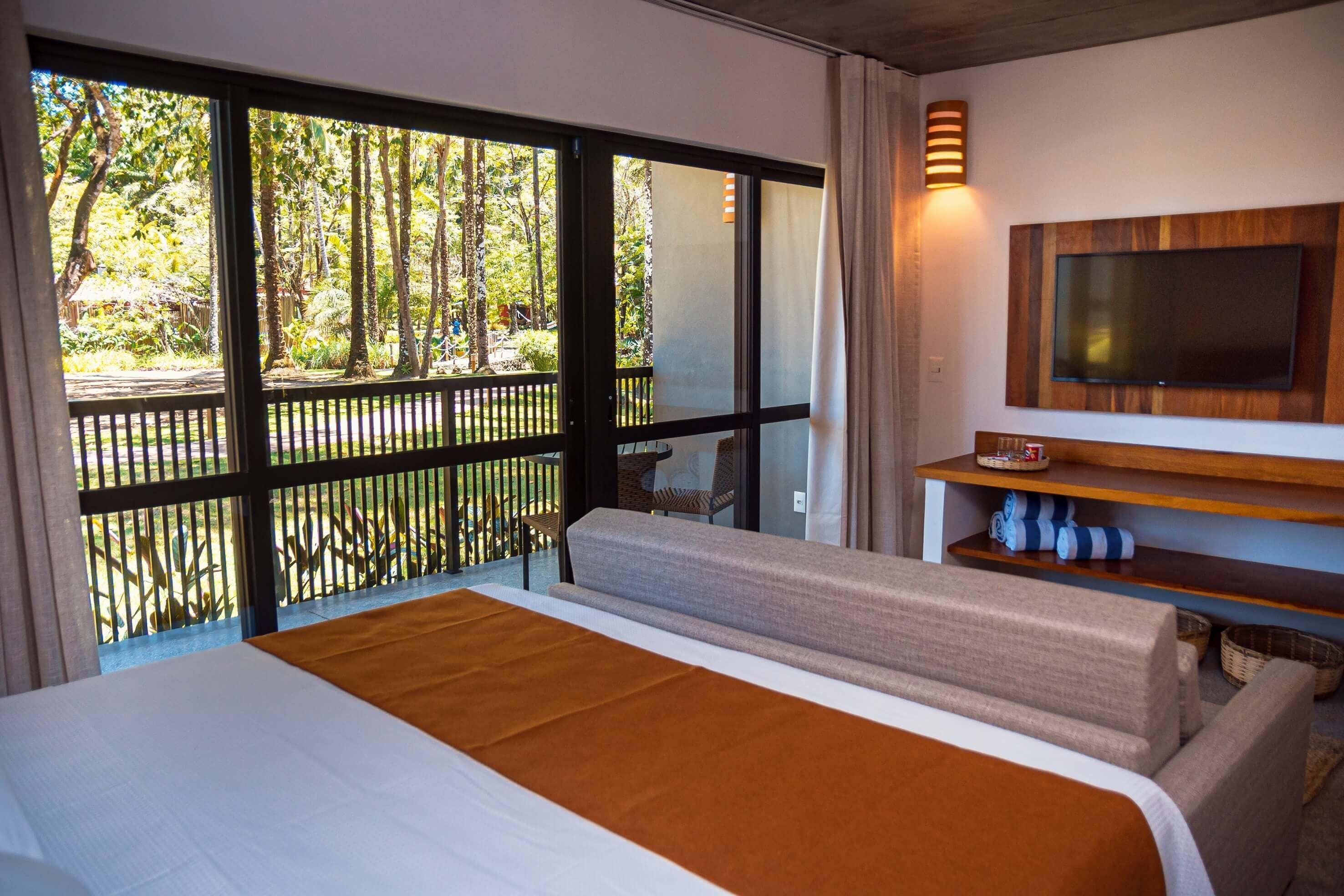 A hotel room with a large bed, a TV mounted on a wooden shelf, and a balcony with a view of trees.