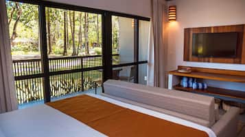A hotel room with a large bed, a TV mounted on a wooden shelf, and a balcony with a view of trees.