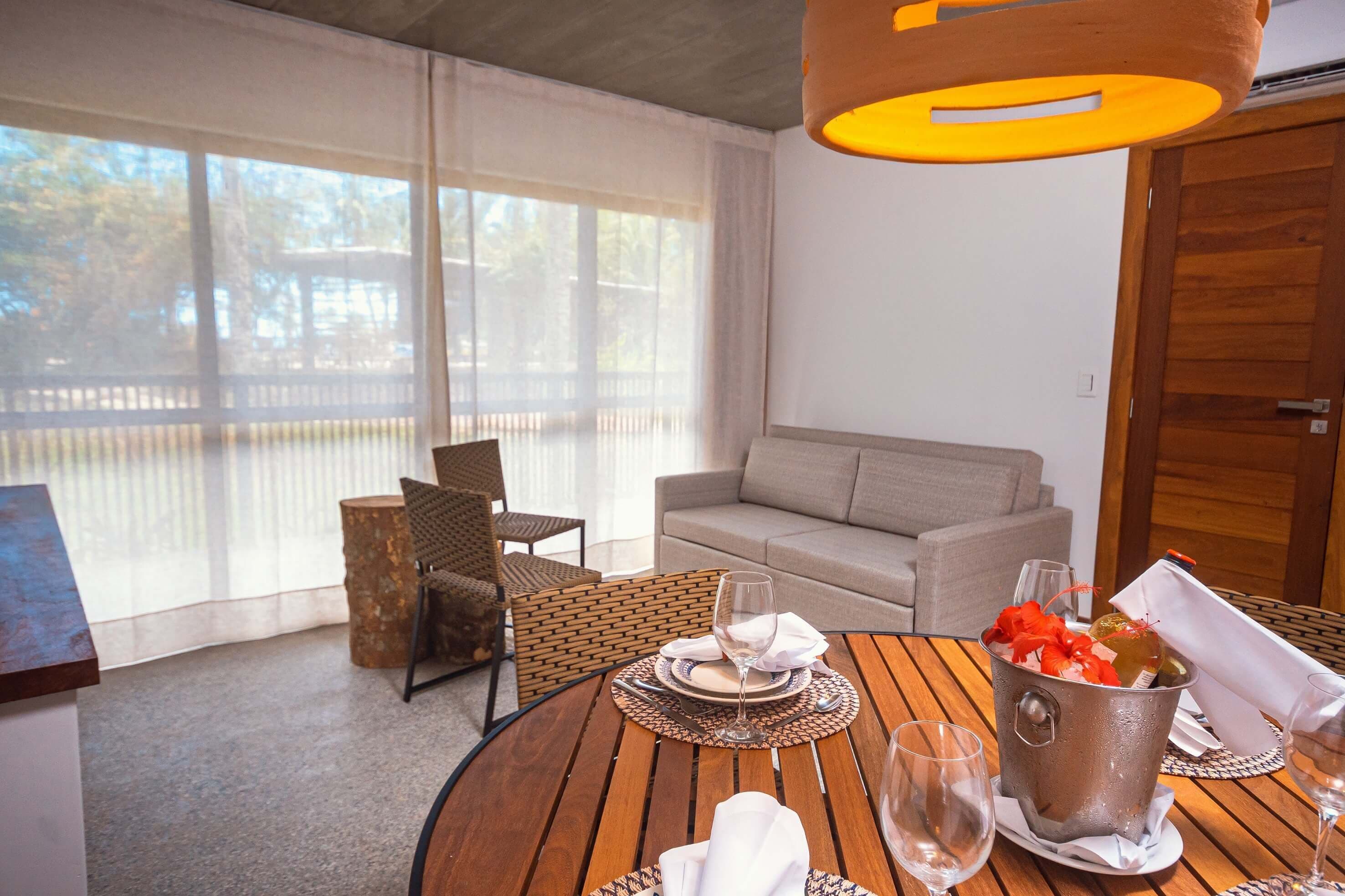 A dining area with a round wooden table set for two, a metal bucket with a bottle and glasses, and a grey sofa in the background.