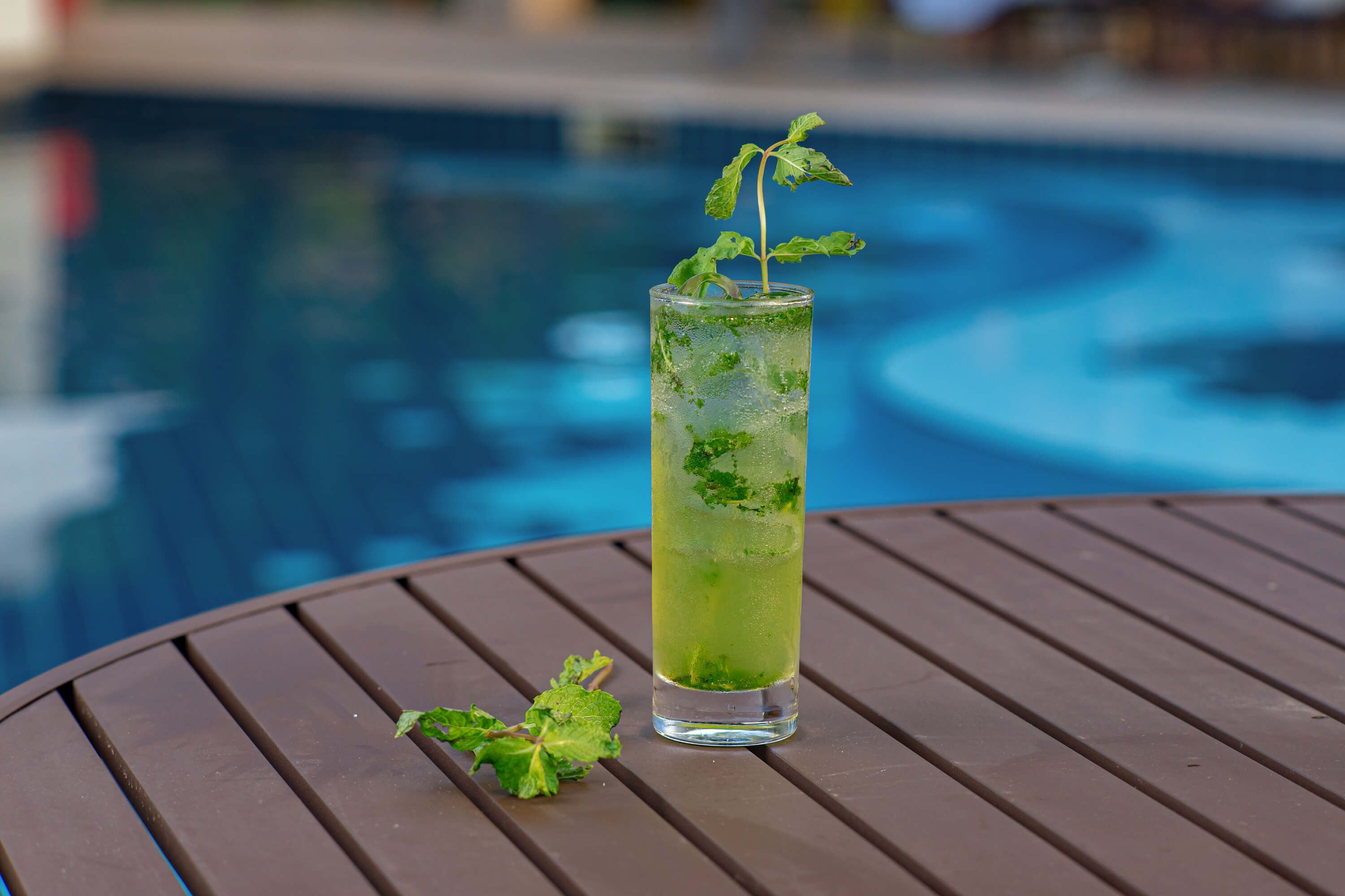 Un vaso alto con bebida verde y hojas de menta sobre una mesa redonda junto a una piscina.